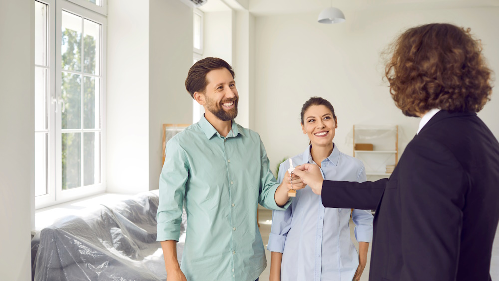 Ein Paar nimmt Schlüssel von einem Immobilienmakler in einem hellen Raum entgegen. Ein Sofa ist mit Plastik abgedeckt.
