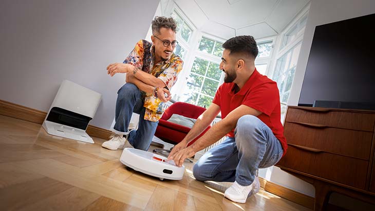 Zwei Männer hocken auf einem Holzboden und betrachten einen weißen Staubsaugerroboter. Im Hintergrund sind Möbel und ein Fenster.