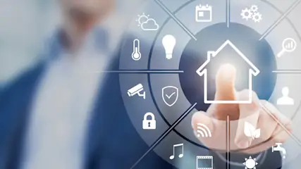 Ein Finger berührt ein Haus-Symbol auf einer runden Oberfläche mit Icons für Wetter, Sicherheit und Medien. Eine Person im Anzug ist unscharf.