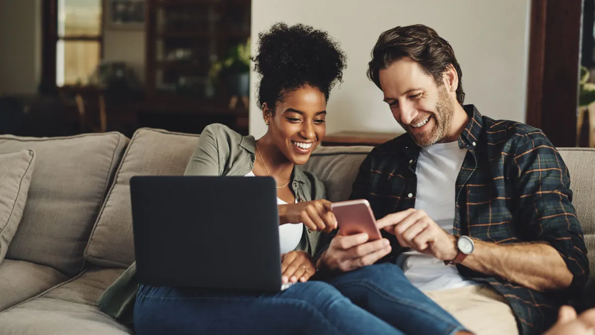 Eine Frau und ein Mann sitzen auf einem Sofa. Sie hat einen Laptop auf dem Schoß, er hat ein Handy in der Hand.