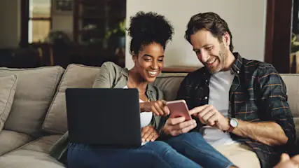 Ein Paar sitzt auf einem Sofa mit Laptop und Handy, beide lächeln und schauen auf das Telefon.