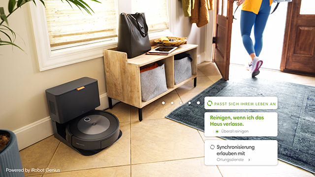 Ein Roboterstaubsauger steht neben einem Holzregal mit einer Tasche. Eine Person verlässt das Haus über eine offene Tür. Eine graue Matte liegt auf dem Boden.