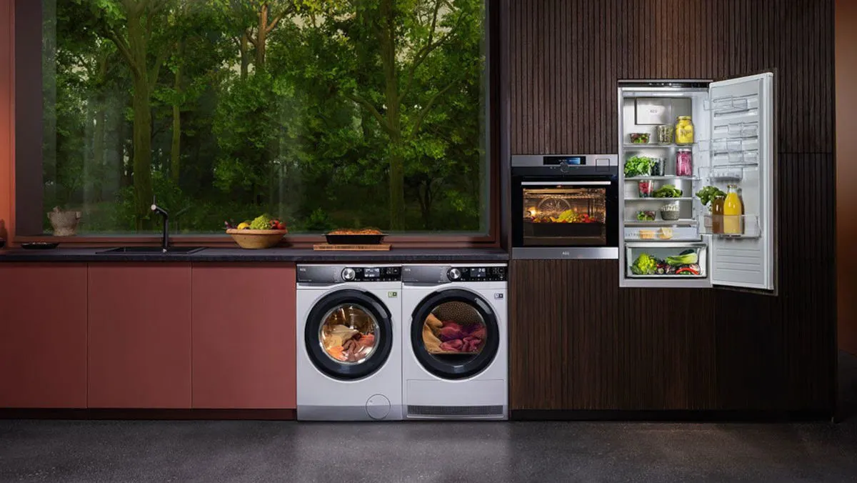 Eine Küche mit Waschmaschine, Trockner, Backofen und offenem Kühlschrank. Draußen ist ein Wald sichtbar.