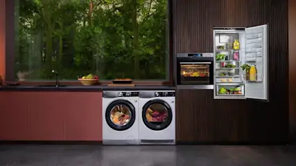 Eine Küche mit Waschmaschine, Trockner, Backofen und offenem Kühlschrank. Draußen ist ein Wald sichtbar.