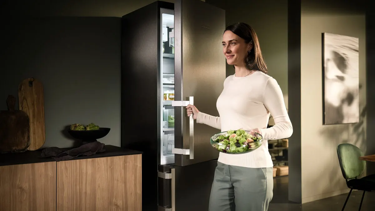 Eine Frau mit Salatschüssel öffnet einen Kühlschrank in einer Küche mit Holzschrank und Stuhl.