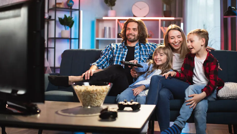 Eine Familie sitzt auf einer blauen Couch und schaut fern. Popcorn und Controller stehen auf dem Tisch vor ihnen.