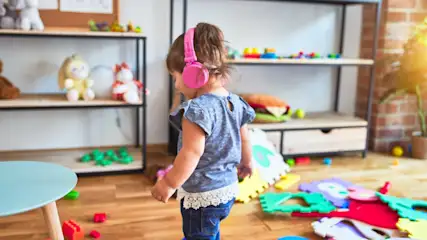Ein Kind mit rosafarbenen Kopfhörern steht in einem Raum mit Spielzeug und Regalen. Puzzlematten liegen auf dem Holzboden.