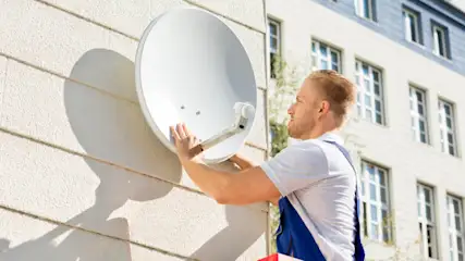 Ein Mann montiert eine weiße Satellitenschüssel an einer hellen Hauswand. Im Hintergrund ist ein Gebäude mit Fenstern zu sehen.