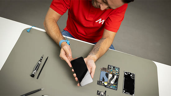 Ein MediaMarkt-Mitarbeiter hält ein auseinander gebautes Smartphone in der Hand während neben ihm Werkzeug liegt.