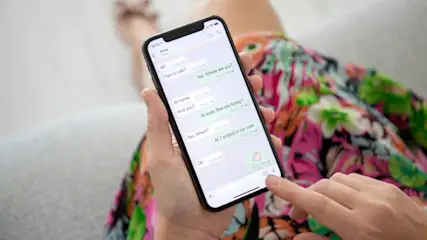 Eine Person hält ein Smartphone mit einer Chat-App in der Hand, während sie auf einer Couch mit einem bunten Kleid sitzt.