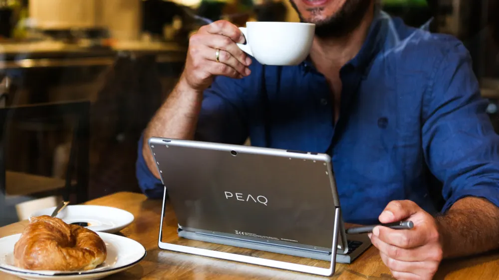 Ein Mann mit blauem Hemd trinkt Kaffee neben einem Tablet mit Stift und einem Croissant auf einem Tisch.