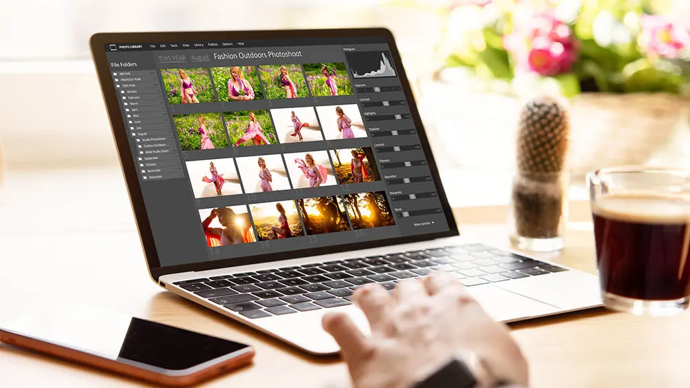 Ein Laptop zeigt Fotoshooting-Bilder, daneben ein Handy, ein Kaktus, ein Glas mit dunklem Getränk und eine Hand auf der Tastatur.