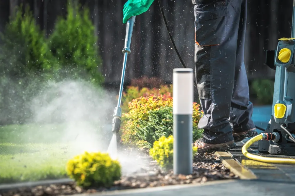 Eine Person mit Arbeitshose und Handschuhen bedient einen Hochdruckreiniger im Garten, umgeben von Pflanzen und Büschen.