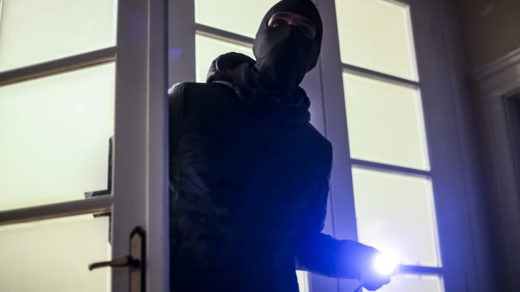 Maskierter Mann mit Taschenlampe in der Hand betritt eine Tür. 
