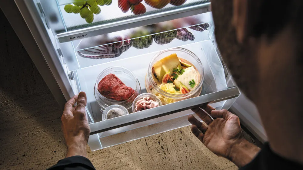 Ein Mann öffnet einen Kühlschrank mit Obst, Gemüse, rohem Fleisch und Käse in durchsichtigen Behältern auf mehreren Regalen.