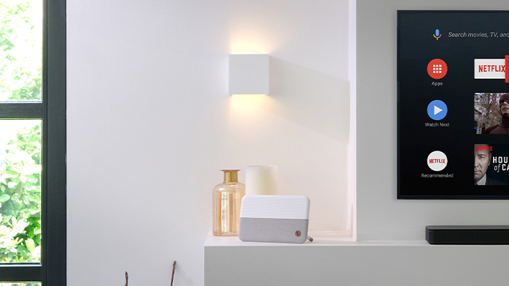 Minimalistisch eingerichteter Raum mit Fenster, Lampe, Flasche, Gerät und Fernseher an einer weißen Wand.