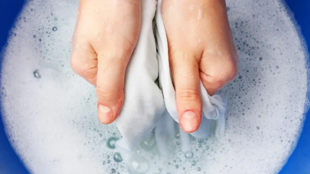 Zwei Hände wringen ein weißes Tuch über einem blauen Behälter voller Seifenwasser mit vielen Blasen.