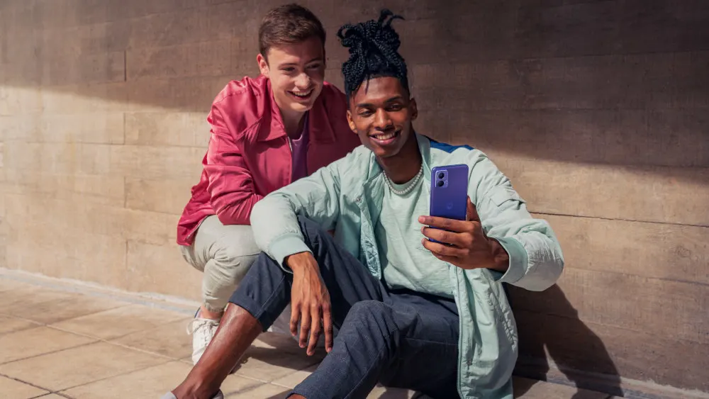 Zwei lächelnde Personen vor einer Wand. Eine Person hält ein blaues Mobiltelefon. Beide tragen Jacken.