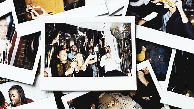 Auf einem Tisch liegen Fotos mit weißen Rändern, die Personen bei einer Party mit Ballons und "Happy"-Schriftzug zeigen.