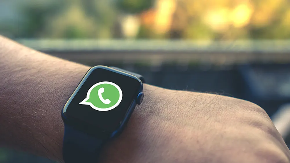 Eine schwarze Smartwatch mit grünem WhatsApp-Symbol wird am Handgelenk getragen, im Hintergrund ist eine unscharfe Natur zu sehen.