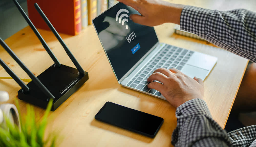 Ein Laptop, ein Smartphone und ein Router stehen auf einem Tisch, ein Mann bedient das Laptop.