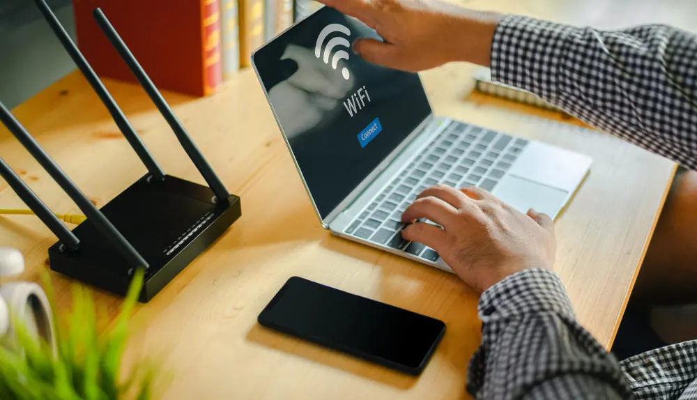 Ein Schreibtisch mit einem schwarzen Router, einem schwarzen Handy und einem Laptop mit einem WLAN-Symbol auf dem Bildschirm. Jemand tippt.