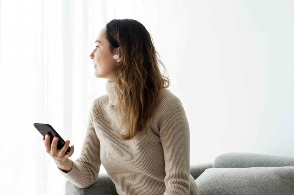 Eine Frau mit einem beigen Rollkragenpullover sitzt auf einem grauen Sofa und hält ein schwarzes Telefon in der Hand. Sie trägt Kopfhörer.