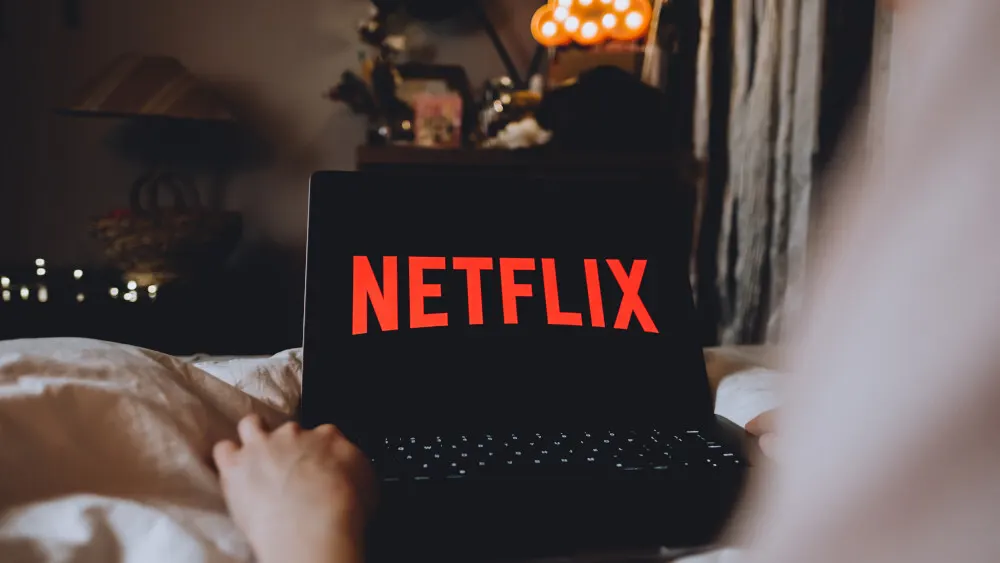 Eine Person im Bett benutzt einen Laptop mit rotem Netflix-Logo auf schwarzem Bildschirm. Lichter und Vorhänge sind im Hintergrund zu sehen.