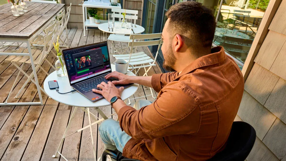 Ein Mann sitzt an einem runden, weißen Tisch im Freien und benutzt einen Laptop, der an eine externe Festplatte angeschlossen ist. Weitere Tische und Stühle sind im Hintergrund zu sehen.