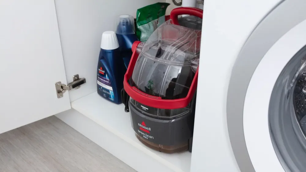Im Schrank neben der Waschmaschine stehen Reinigungsflüssigkeiten und ein tragbarer Fleckenreiniger mit rotem Griff.
