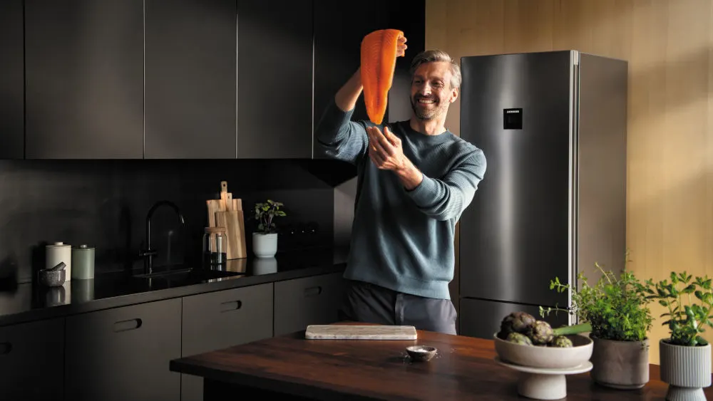 In einer Küche hält ein Mann eine orangefarbene Silikonform hoch. Im Hintergrund befinden sich Schränke, ein Kühlschrank und Pflanzen.