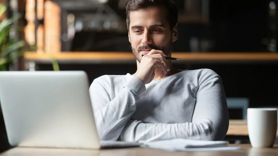 Ein Mann sitzt an einem Tisch mit Laptop und Stift in der Hand, daneben eine Tasse; Pflanzen im Hintergrund.