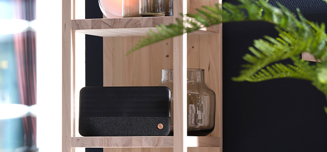 Ein Holzregal mit einer schwarzen Box, einer braunen Vase und einer Kerze, dekoriert mit grünen Pflanzen vor einem dunklen Hintergrund.