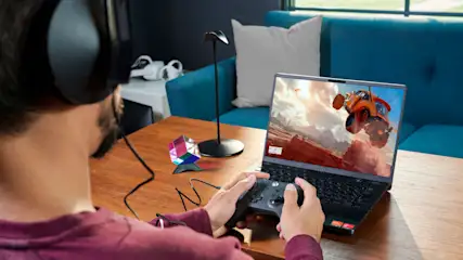 Mann mit Kopfhörern spielt mit Controller ein Rennspiel auf einem Laptop. Ein Rubik-Würfel und eine Lampe stehen auf dem Tisch.