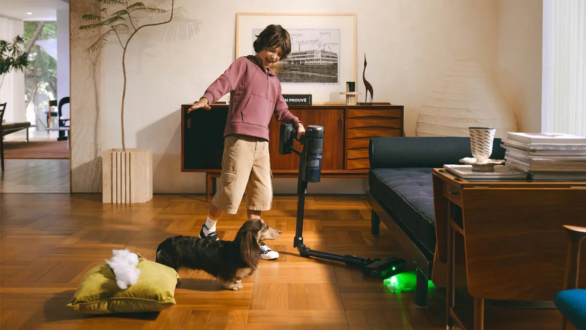 Ein Kind saugt mit dem Roborock H60 Ultra unter einem Sofa, daneben steht ein kleiner Hund.