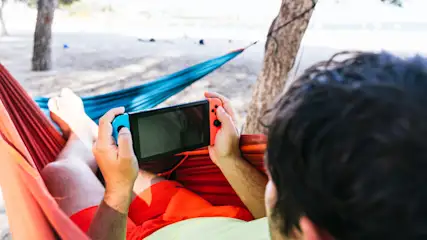 Mann liegt in einer Hängematte und spielt auf einer Nintendo Switch mit blauem und rotem Controller.