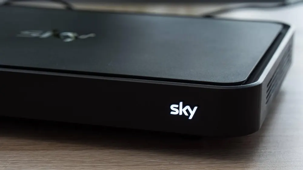 Ein schwarzer Sky-Receiver steht auf einem hellen Holztisch. Das Sky-Logo ist in weißer Schrift auf der Vorderseite des Geräts sichtbar.