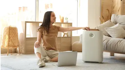 Eine Frau sitzt auf einem Teppich mit Laptop, neben einem Luftreiniger, vor einem Sofa und einem Regal in einem hellen Raum.