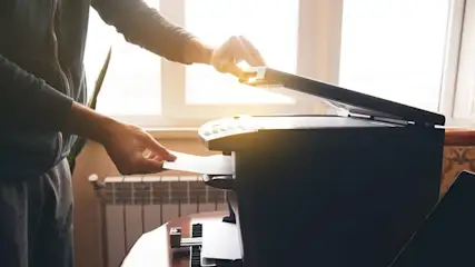 Eine Person in Jacke und grauer Hose bedient einen schwarzen Drucker, indem sie Papier einlegt. Im Hintergrund sind ein Fenster und ein Heizkörper sichtbar.