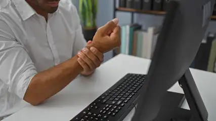 Mann in weißem Hemd hält Handgelenk vor Computer und schwarzer Tastatur auf weißem Tisch.