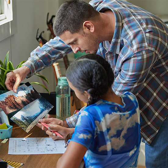 Vater und Kind betrachten Fotos am Tisch. Das Kind hält einen Stift über einem Blatt Papier mit Zeichnungen. Eine Wasserflasche steht daneben.