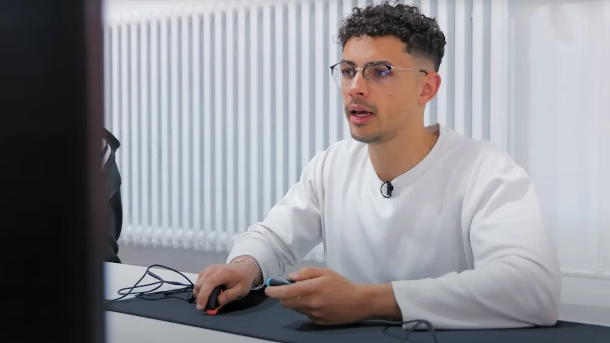Ein Mann mit Brille und weißem Pullover sitzt an einem Tisch mit Maus und Mikrofon vor einem Monitor in einem hellen Raum.