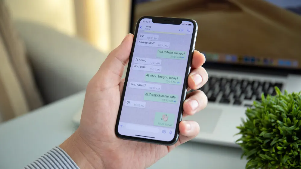 Hand hält ein Smartphone mit einer Chat-App. Auf dem Display sind Textnachrichten zu sehen, im Hintergrund ein Laptop und eine Pflanze.
