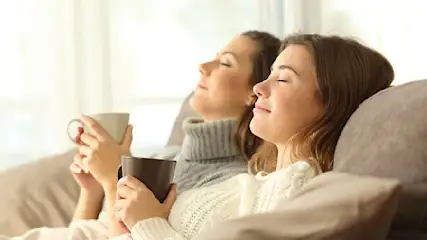 Zwei Frauen entspannen auf einem Sofa mit Tassen in der Hand, die Augen geschlossen, vor einem hellen Fenster.