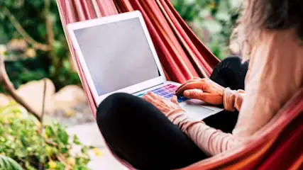 Eine Person sitzt in einer rot gestreiften Hängematte und benutzt einen Laptop mit bunten Tasten. Im Hintergrund sind grüne Pflanzen.