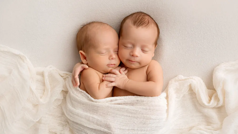 Zwei schlafende Babys liegen eng aneinander in einem weißen Tuch auf einem hellen Hintergrund.