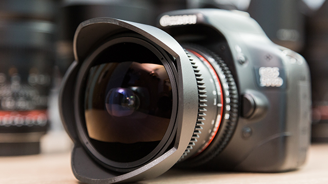Eine schwarze Kamera mit einem großen Objektiv steht auf einem Tisch. Das Objektiv hat einen grauen Ring und eine Gegenlichtblende.