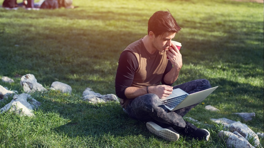 Ein Mann sitzt mit gekreuzten Beinen im Gras, arbeitet am Laptop und hält einen roten Becher in der Hand. Steine liegen im Gras.