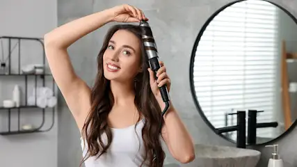 Eine Frau mit lange braunen Haar stylt sich Locken mit einem Lockenstab
