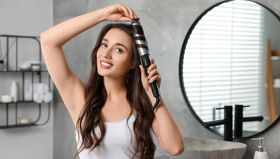 Eine Frau mit langen braunen Haaren stylt sich Locken mit einem Lockenstab.